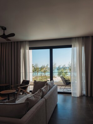 Ein modernes Wohnzimmer mit einer Couch, Sesseln und einem Tisch. Ein Deckenventilator ist sichtbar, und das Zimmer hat große Fenster mit durchscheinenden Vorhängen.