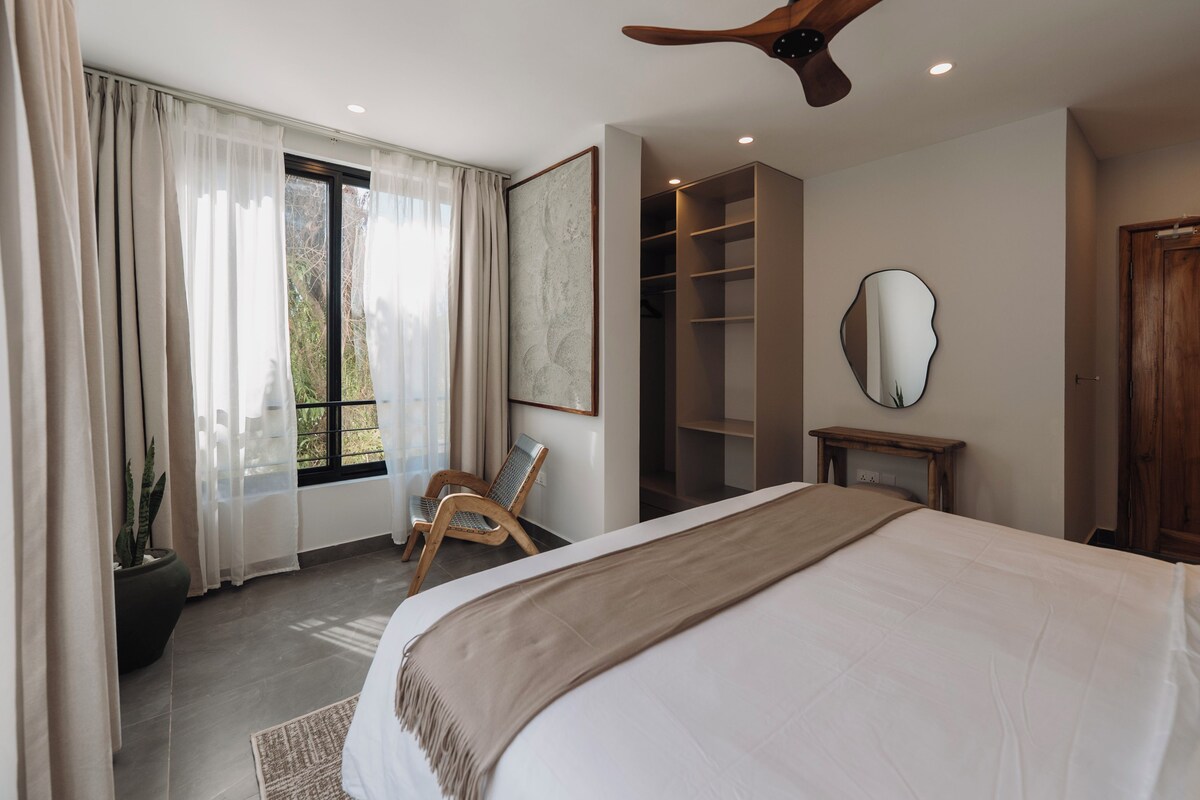 Un dormitorio con una cama grande, un ventilador de techo, una ventana con cortinas translúcidas, una silla, un armario empotrado y un espejo en la pared.