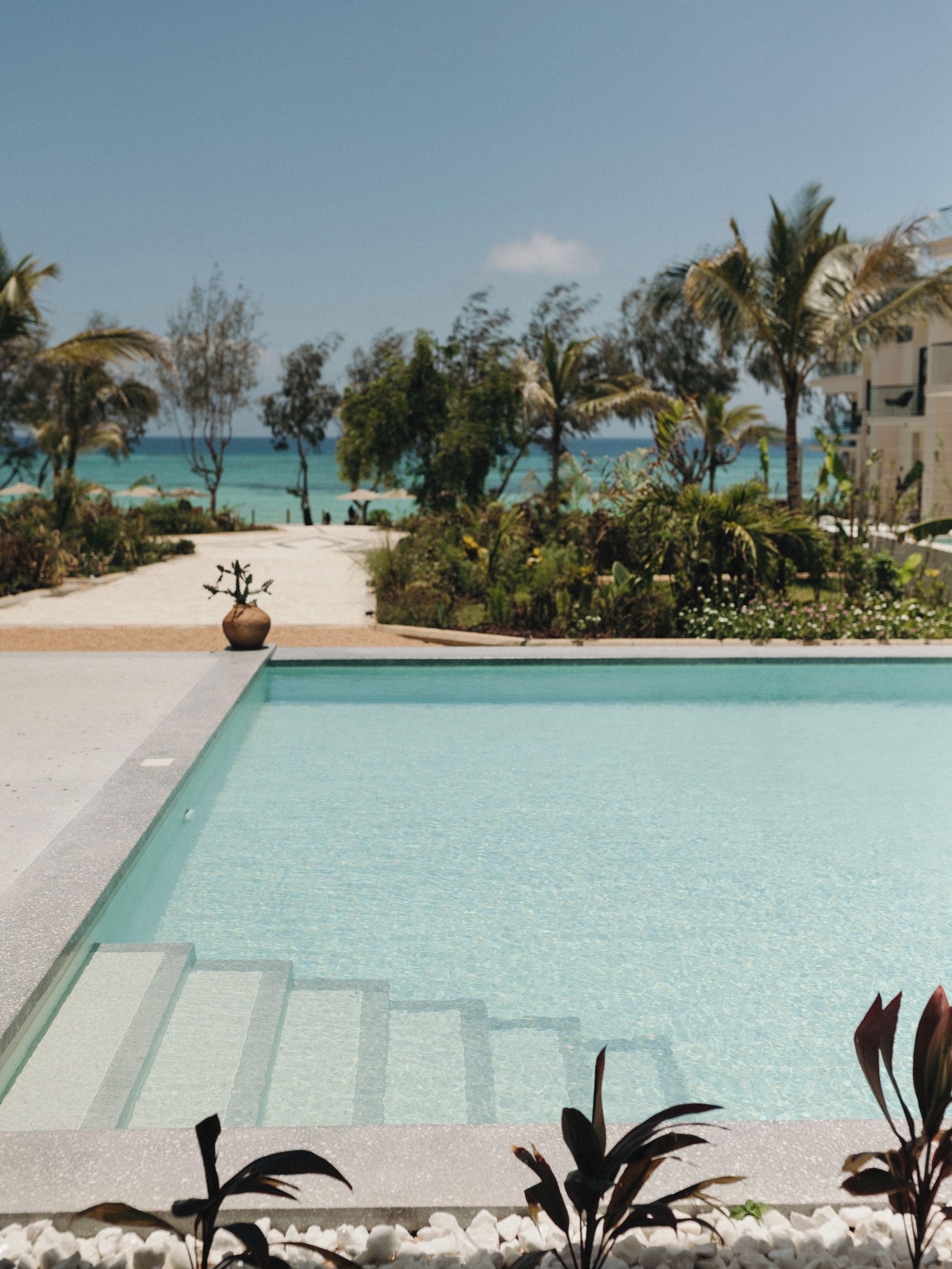 A swimming pool with clear blue water, surrounded by tropical plants and palm trees, leading to a beach with the ocean in the background.