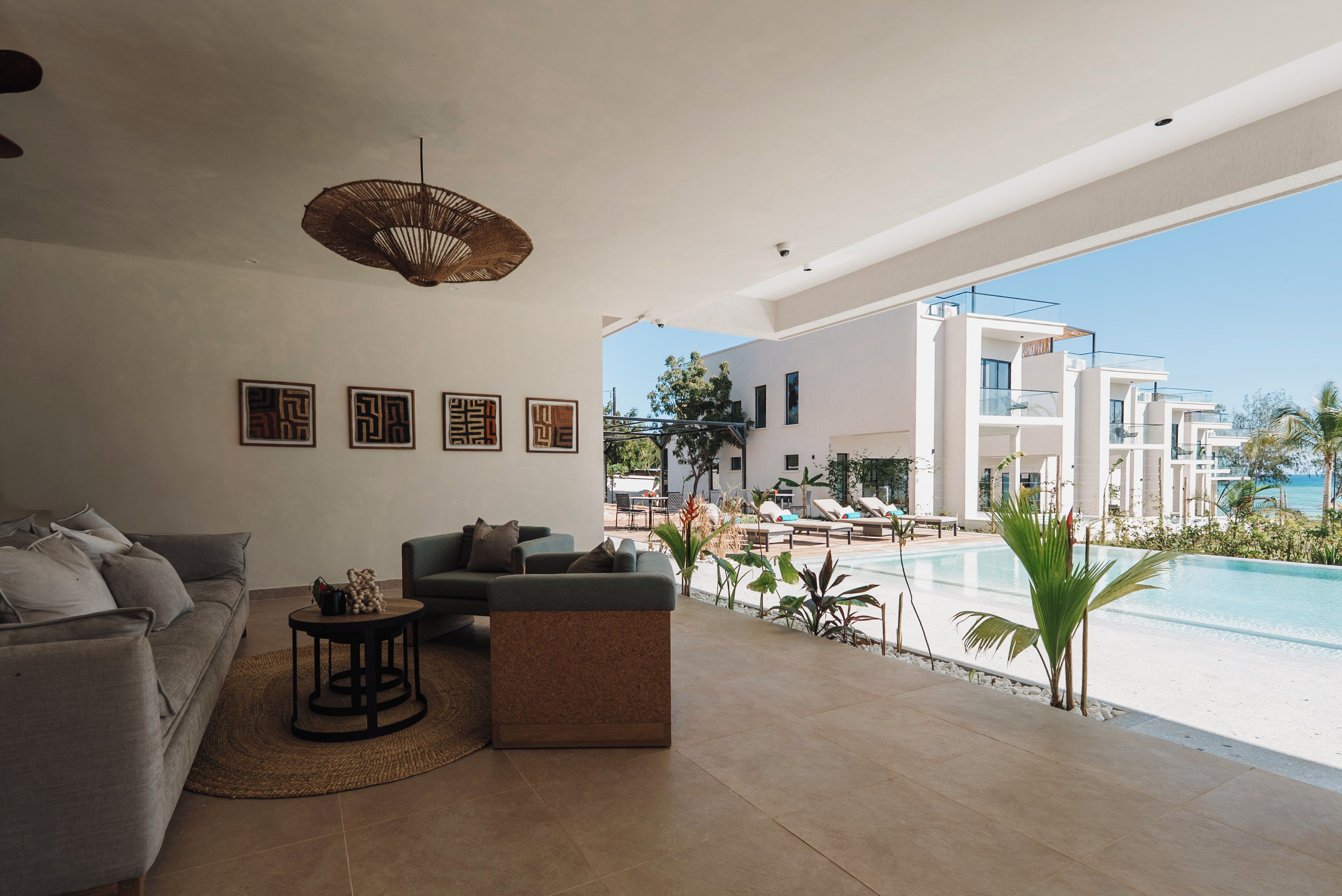 Uma sala de estar moderna com sofá, mesa de centro e luminária pendente. É visível uma área de piscina através das grandes janelas.