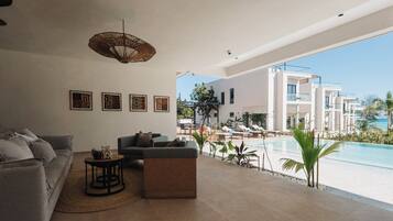 Uma sala de estar moderna com sofá, mesa de centro e luminária pendente. É visível uma área de piscina através das grandes janelas.