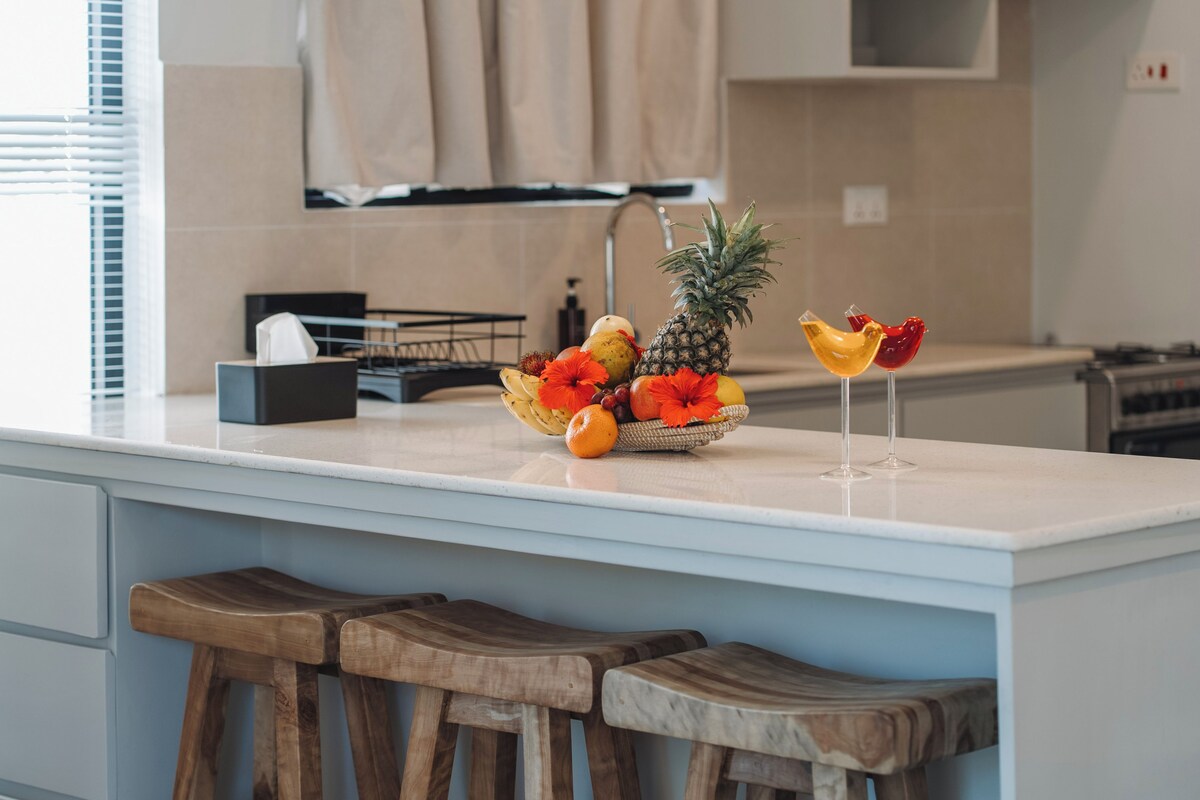 Una encimera de cocina con un frutero, dos copas de vino y taburetes de madera.
