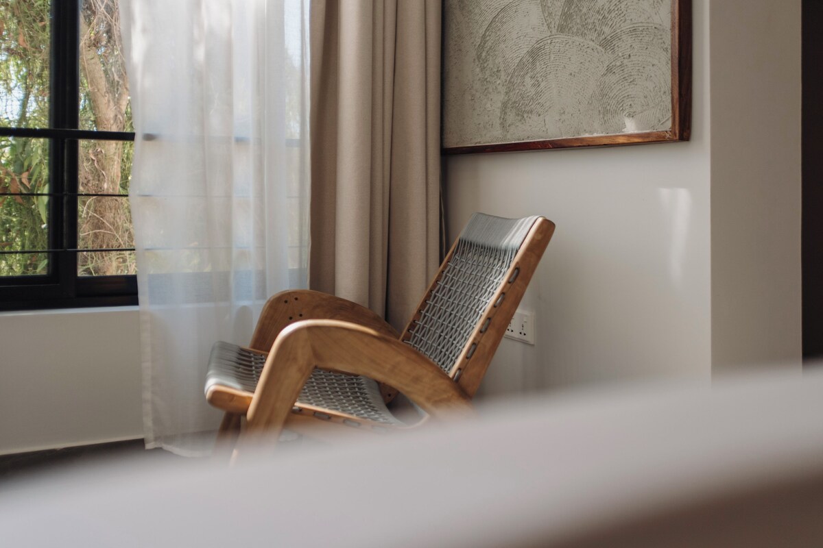 Una habitación con una silla de madera, un radiador, una ventana con cortinas traslúcidas y una obra enmarcada en la pared.