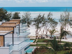 Un hotel frente a la playa con varios balcones con vistas al océano, zona de piscina y jardines tropicales.