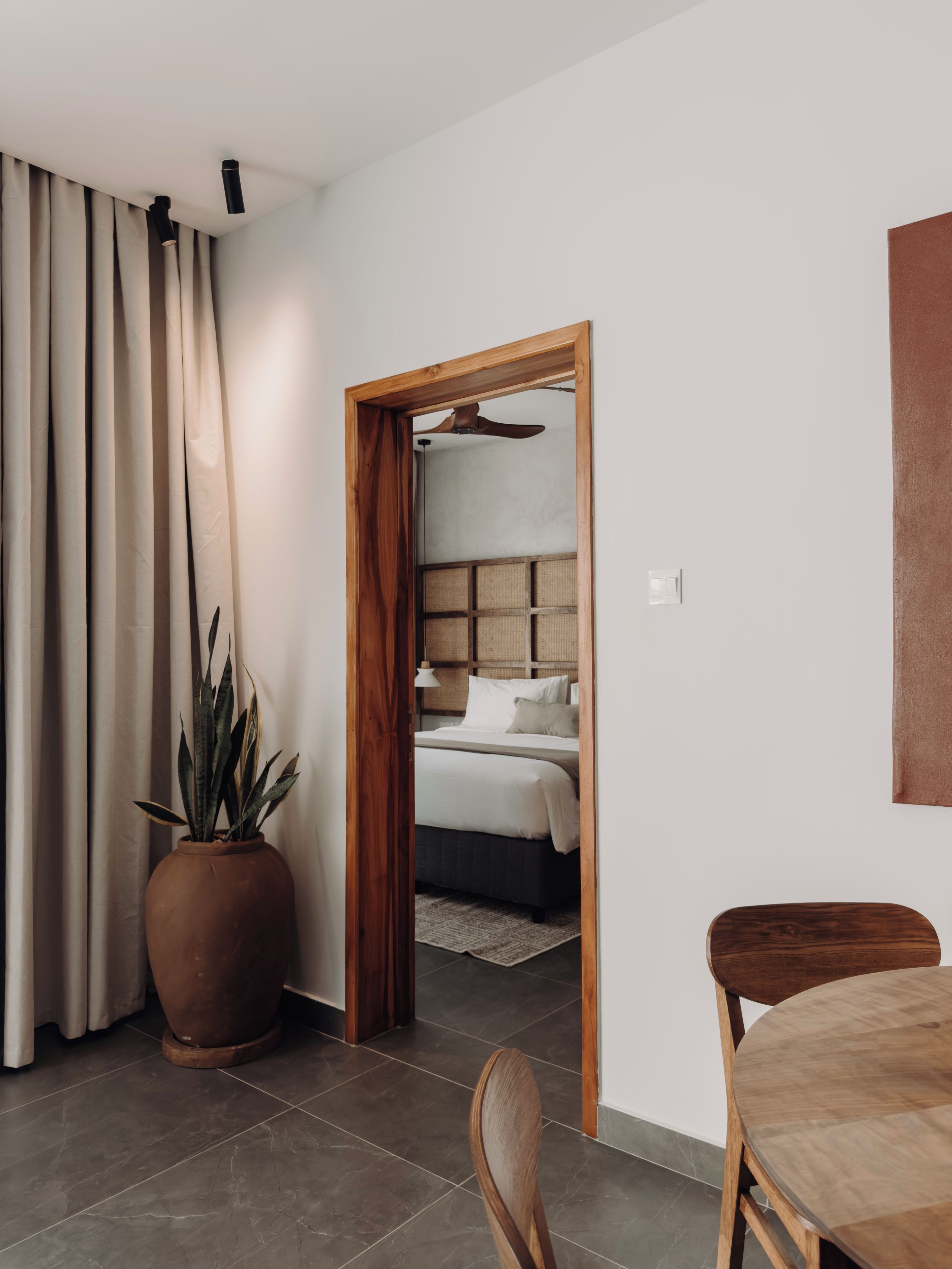 Quarto de hotel moderno com uma cama grande, porta de madeira que dá para um banheiro, uma planta em vaso e um espaço de refeições com mesa e cadeiras.
