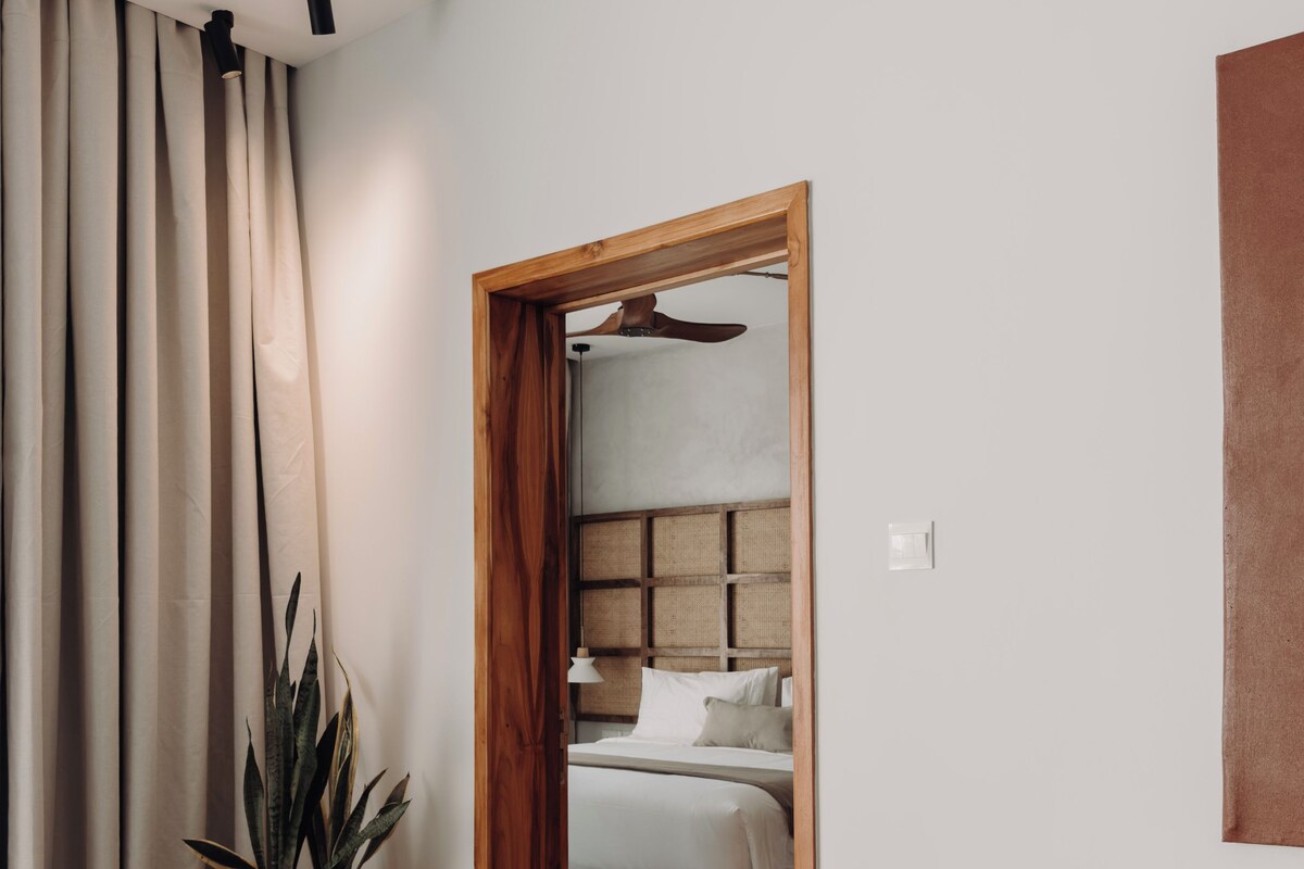 Una habitación de hotel moderna con una cama grande, una puerta de madera que da al baño, una planta en maceta y una zona de comedor con una mesa y sillas.