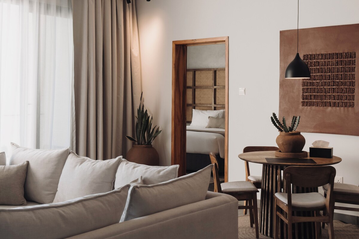 Una sala moderna con un sofá, una mesa de comedor con sillas, una planta en maceta y una lámpara fijada a la pared.