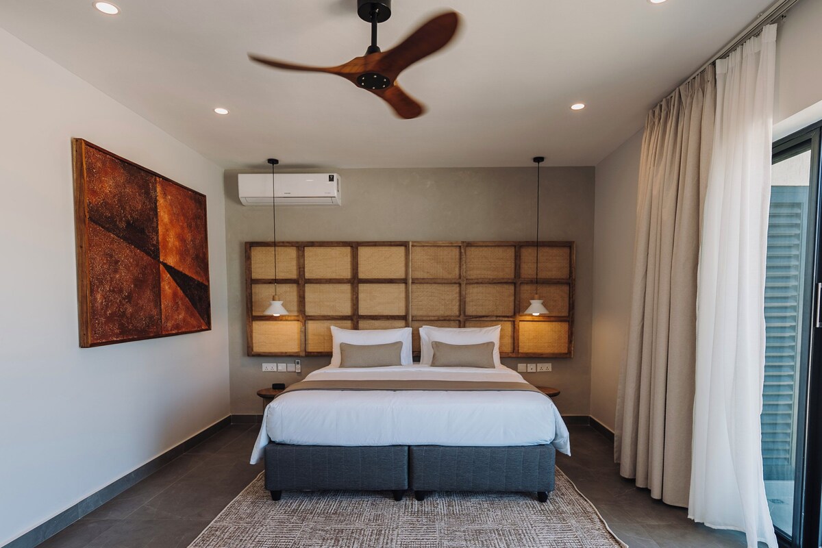 Una habitación de hotel moderna con una cama grande, un ventilador de techo, aire acondicionado instalado en la pared y un amplio ventanal con cortinas.