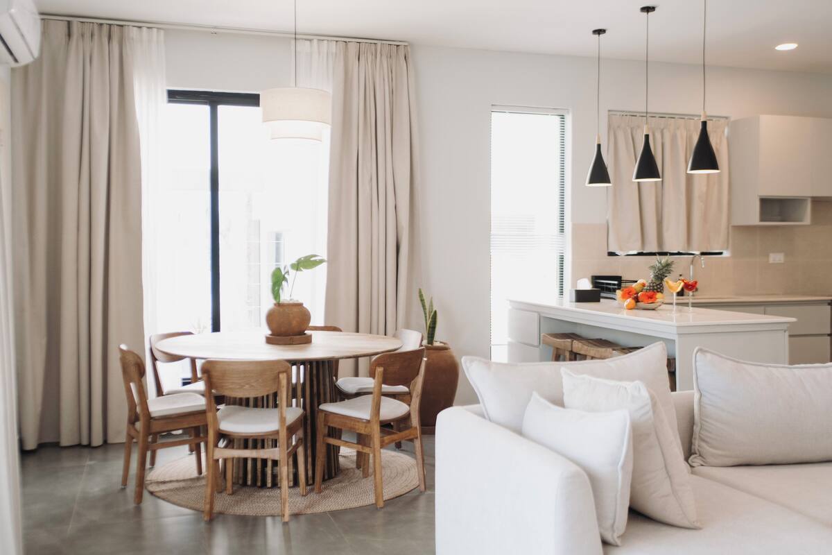 Un salon moderne avec une table de salle à manger ronde, des chaises en bois, un canapé blanc et un coin cuisine doté de luminaires suspendus.