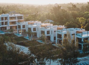 Un hotel moderno de varios pisos con múltiples balcones, piscina y abundante vegetación alrededor.