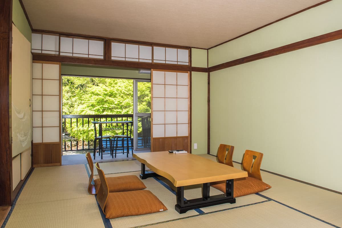 A tatami mat room with a low table and chairs, a sliding door leading to an outdoor area with a view of greenery.