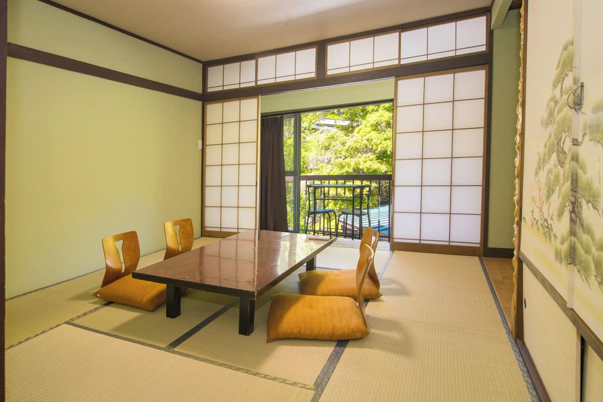 A tatami mat room with a low table and chairs, a sliding door leading to a balcony, and a traditional Japanese painting on the wall.
