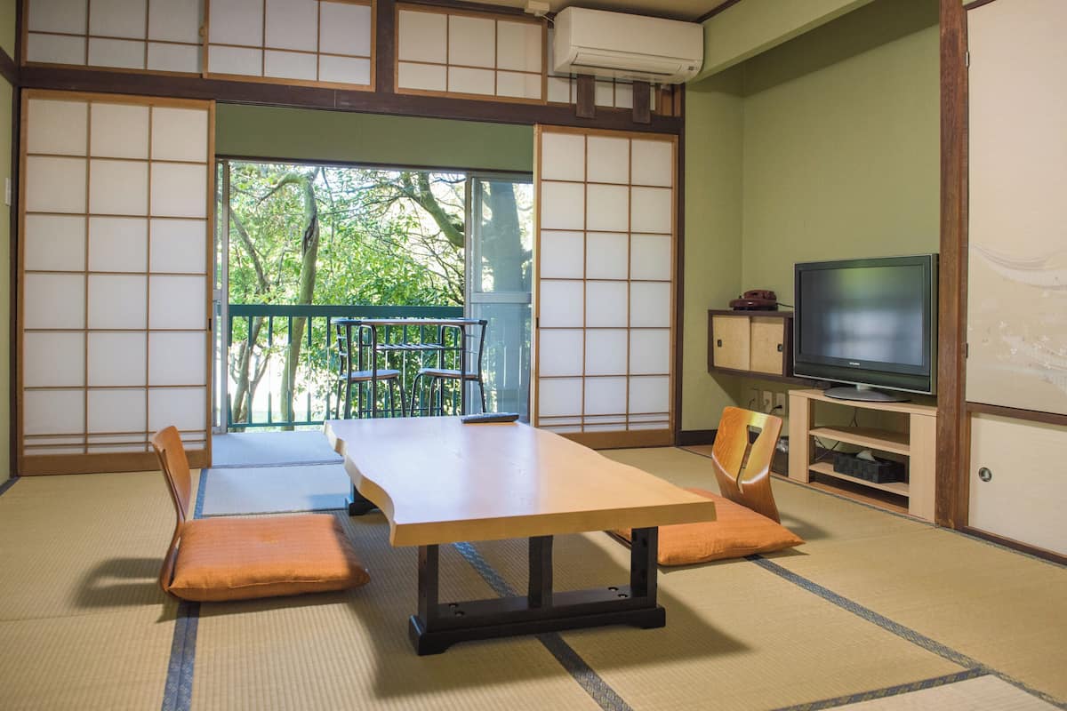 A traditional Japanese room with tatami flooring, a low table with cushions, a television on a cabinet, and a sliding door leading to a balcony with a view of trees.