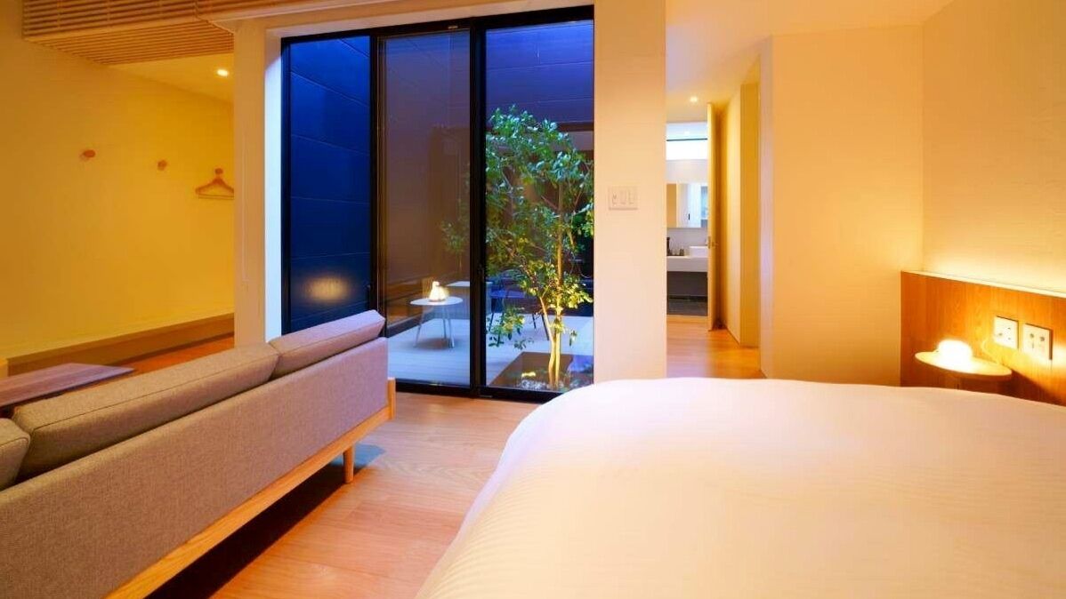A modern hotel room with a large bed, a sofa, a sliding glass door leading to a balcony, and a small table with a lamp.