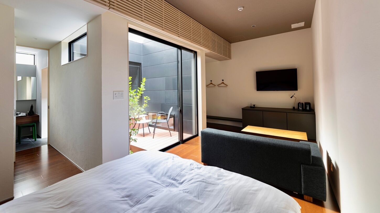 A modern hotel room with a large bed, a TV, a balcony with a dining area, and a sliding door to a patio.