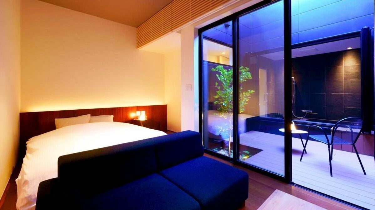 A modern hotel room with a large bed, a sofa, a glass door leading to a balcony with a chair, and a nightstand with a lamp.