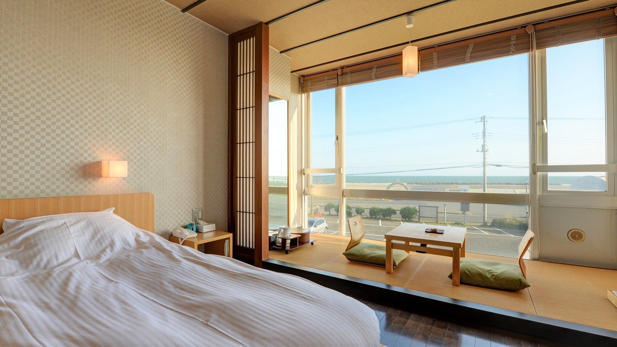A hotel room with a large bed, a small table with a view, and a window overlooking a road and sea.