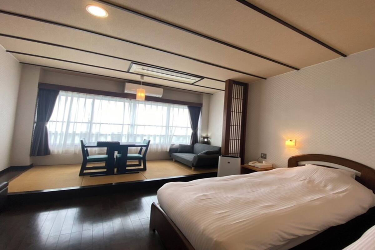 Habitación de hotel con cama, escritorio con silla, sofá y una ventana grande con cortinas.