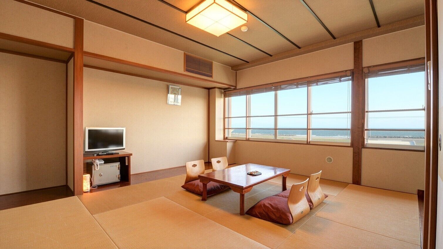 A traditional Japanese room with tatami flooring, a television, a table with cushions, and a large window with a view.