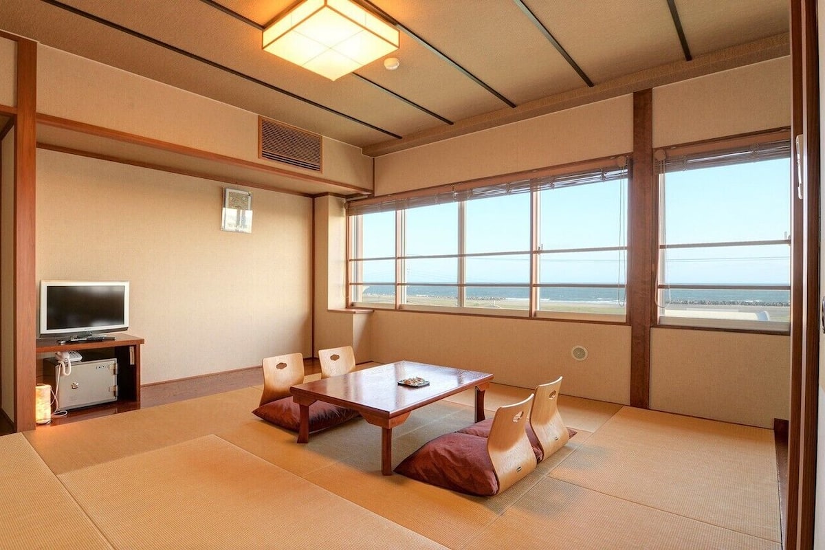 A traditional Japanese room with tatami flooring, a television, a table with cushions, and a large window with a view.