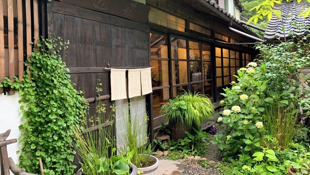 Un edificio japonés tradicional con fachada de madera, ventanas amplias y un jardín con diversas plantas y un sendero de piedra.