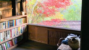 Una habitación con un ventanal que da a un bosque con follaje otoñal. En el interior, hay una estantería llena de libros y una mesita con un jarrón.