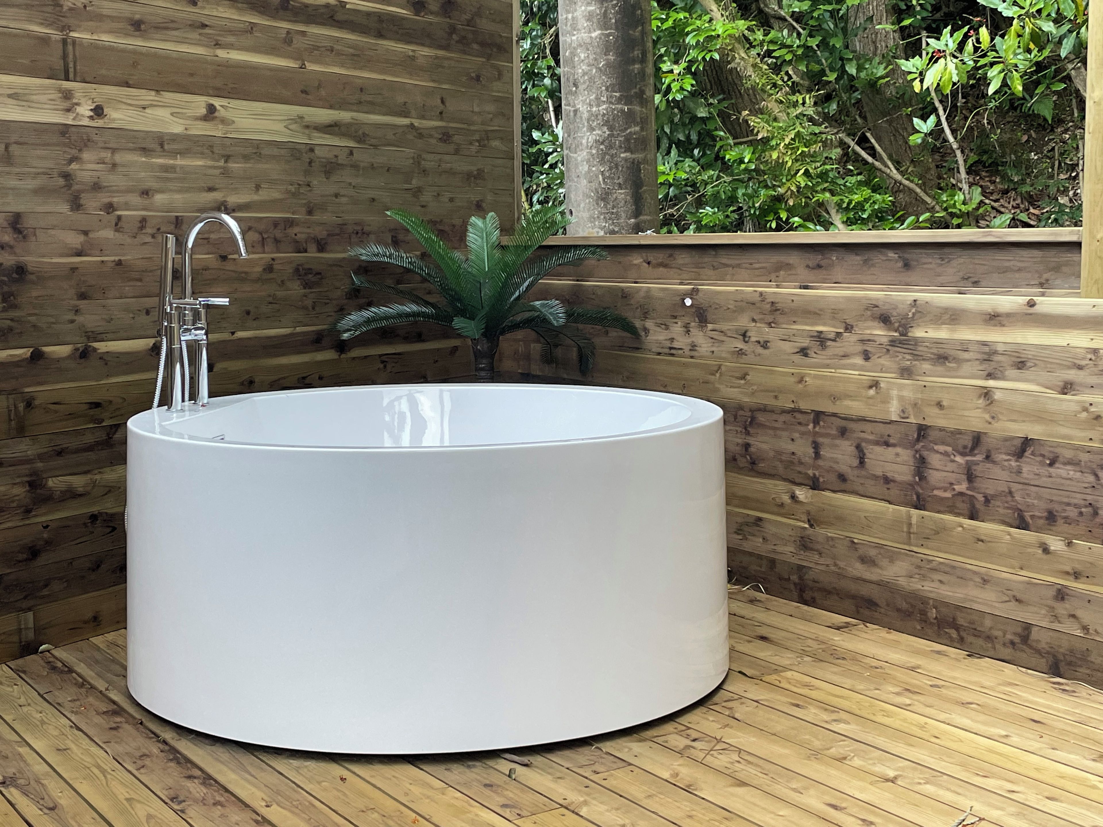 A modern, round, white bathtub in a wooden room with a wooden wall and a window overlooking greenery.