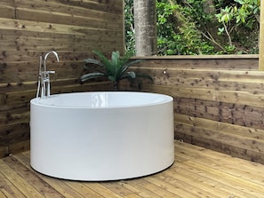 A modern, round, white bathtub in a wooden room with a wooden wall and a window overlooking greenery.