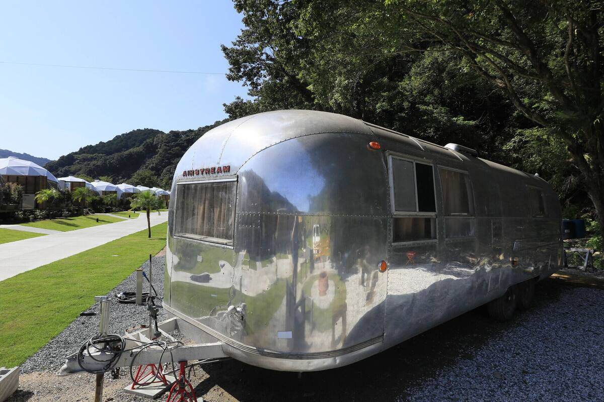 A vintage trailer with a reflective surface, parked on a gravel surface with a grassy area and trees in the background.