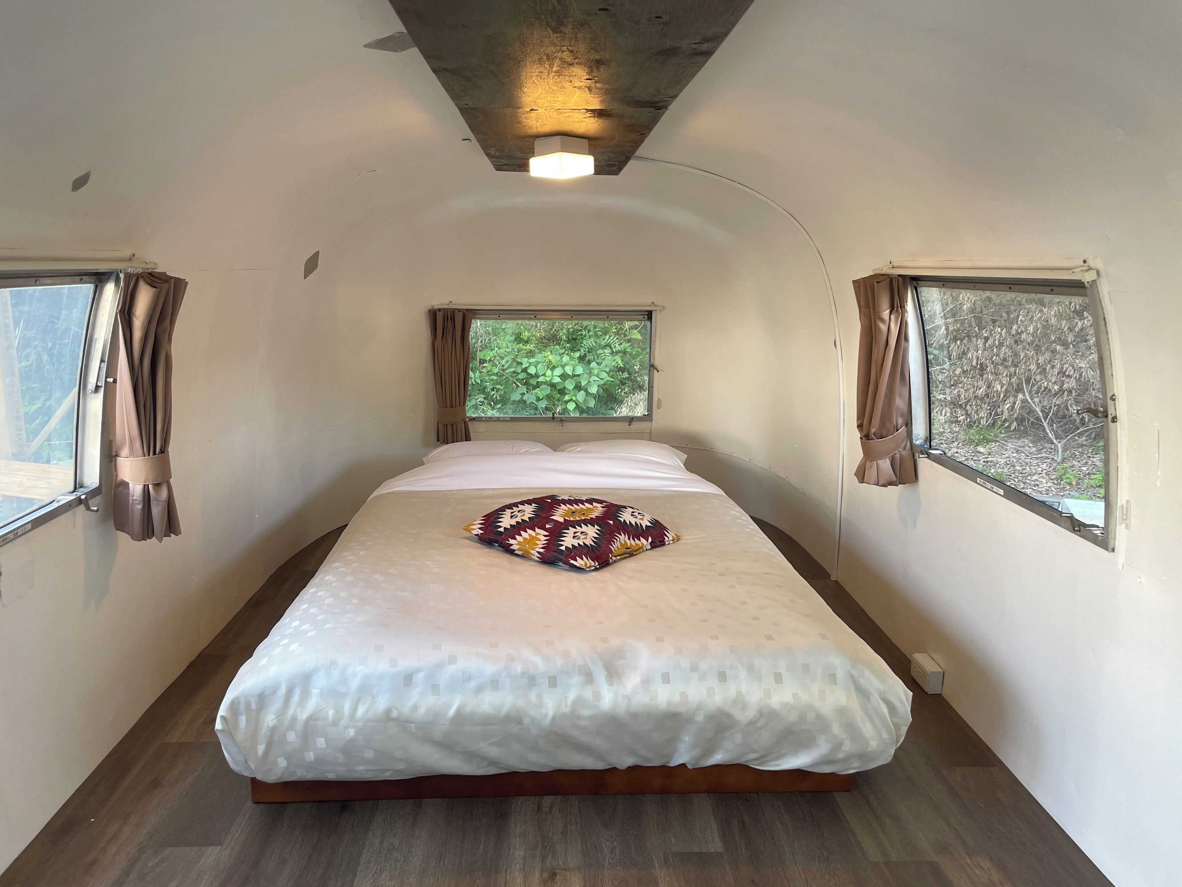 Una habitación estrecha con cama, dos ventanas y una lámpara en el techo.