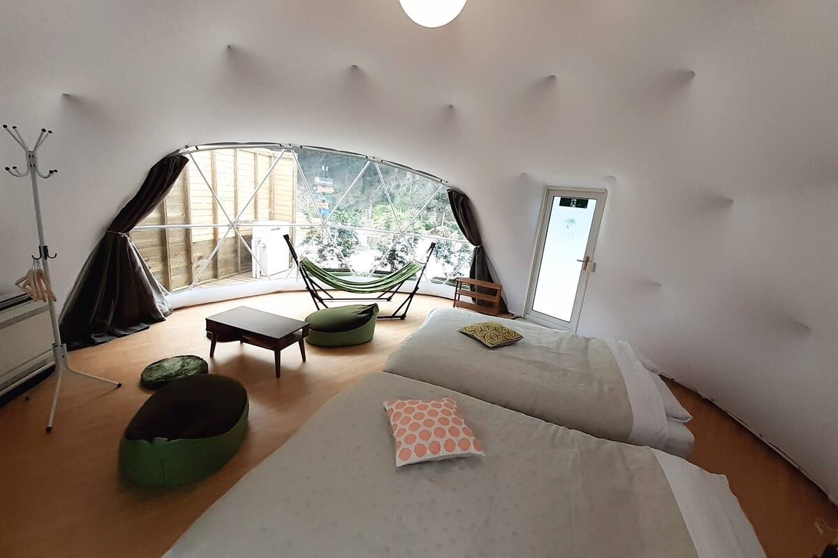 A cozy, circular room with a bed, a chair, a hammock, and a view of greenery outside.