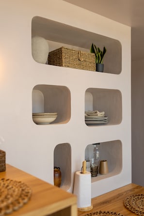 A wall with built-in shelves containing a woven basket, a potted plant, folded plates, a glass, and a roll of paper towels. - Oasis Village (Eforie)