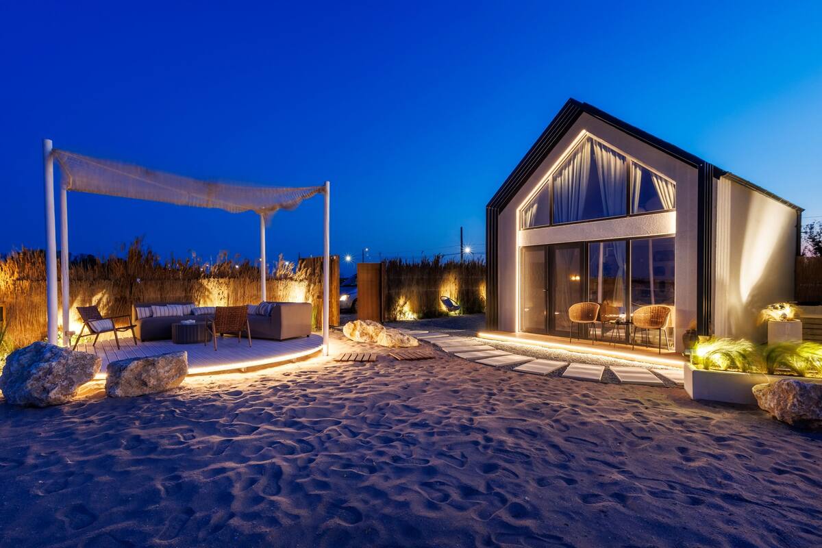A modern beach house with a large glass facade, outdoor seating area, and a well-lit patio.