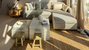 A modern living room with a sectional sofa, a coffee table, and a small wooden stool. There is a large window with curtains, a wall-mounted light, and a hanging light fixture. - Oasis Village (Eforie)
