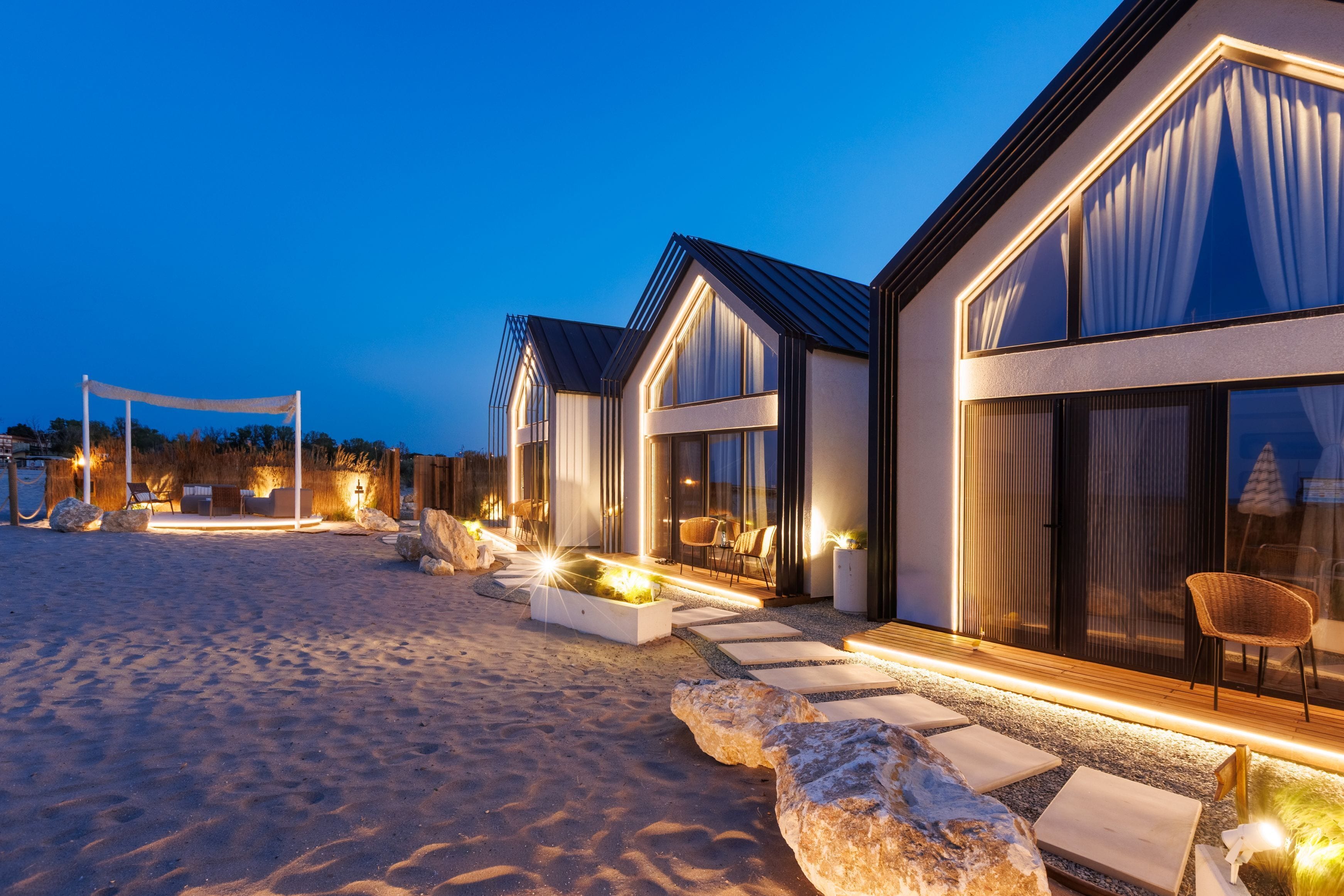 A beachfront hotel with modern architecture, featuring large windows, outdoor seating areas, and a well-lit entrance.