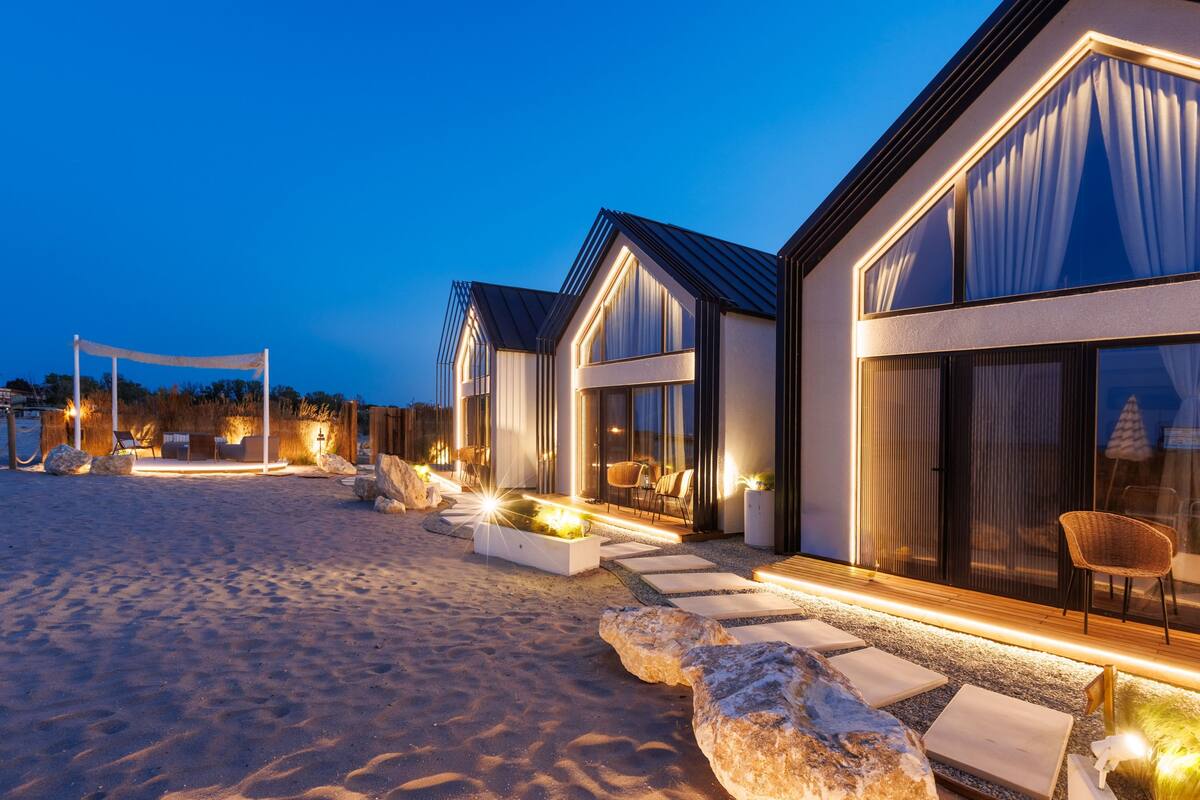 A beachfront hotel with modern architecture, featuring large windows, outdoor seating areas, and a well-lit entrance.