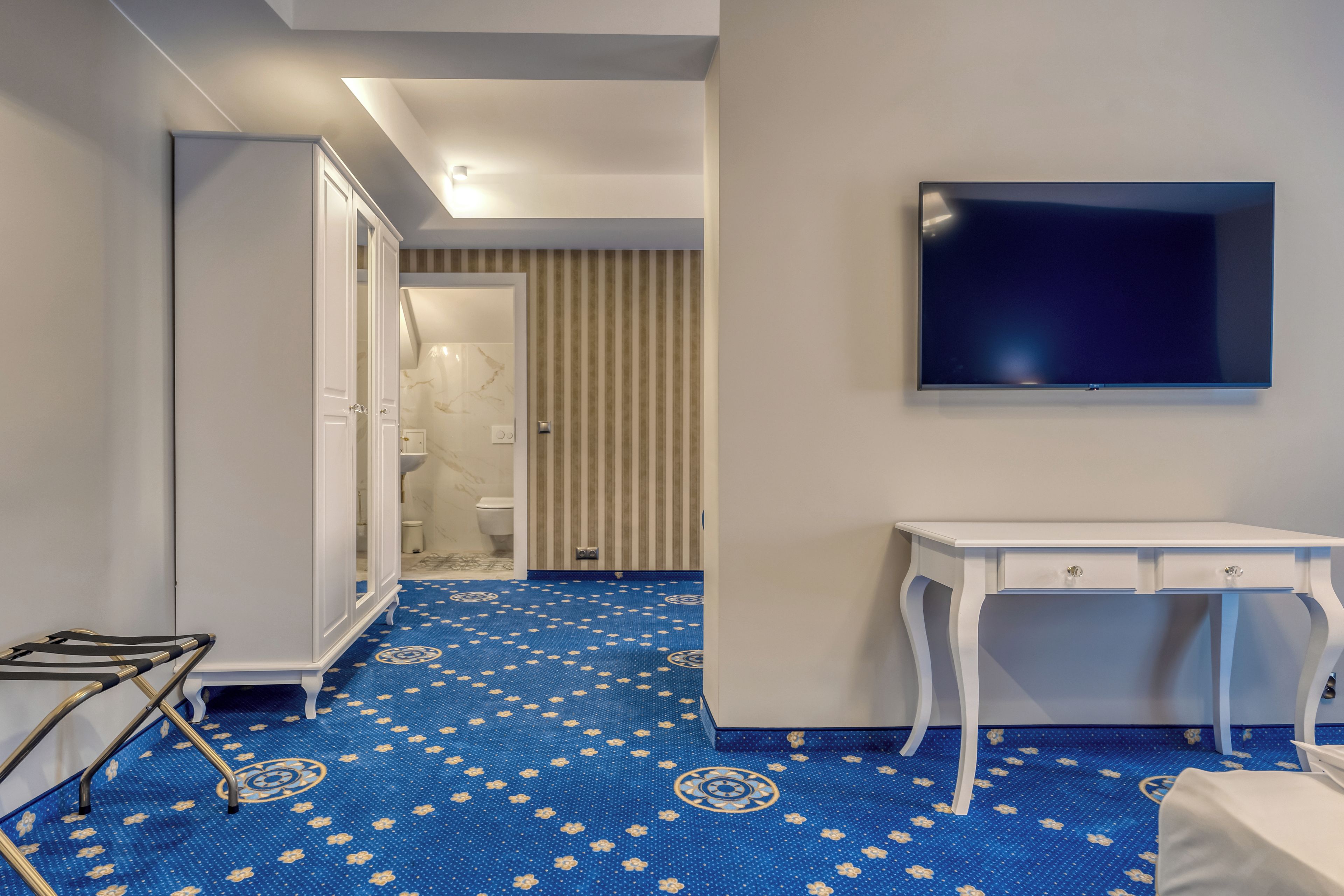 A hotel room with a blue patterned floor, a white desk, a television on the wall, and a wardrobe.