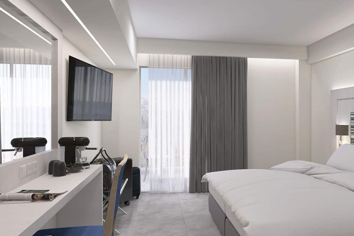 A modern hotel room with a bed, a desk with a computer, a TV, and a window with curtains.