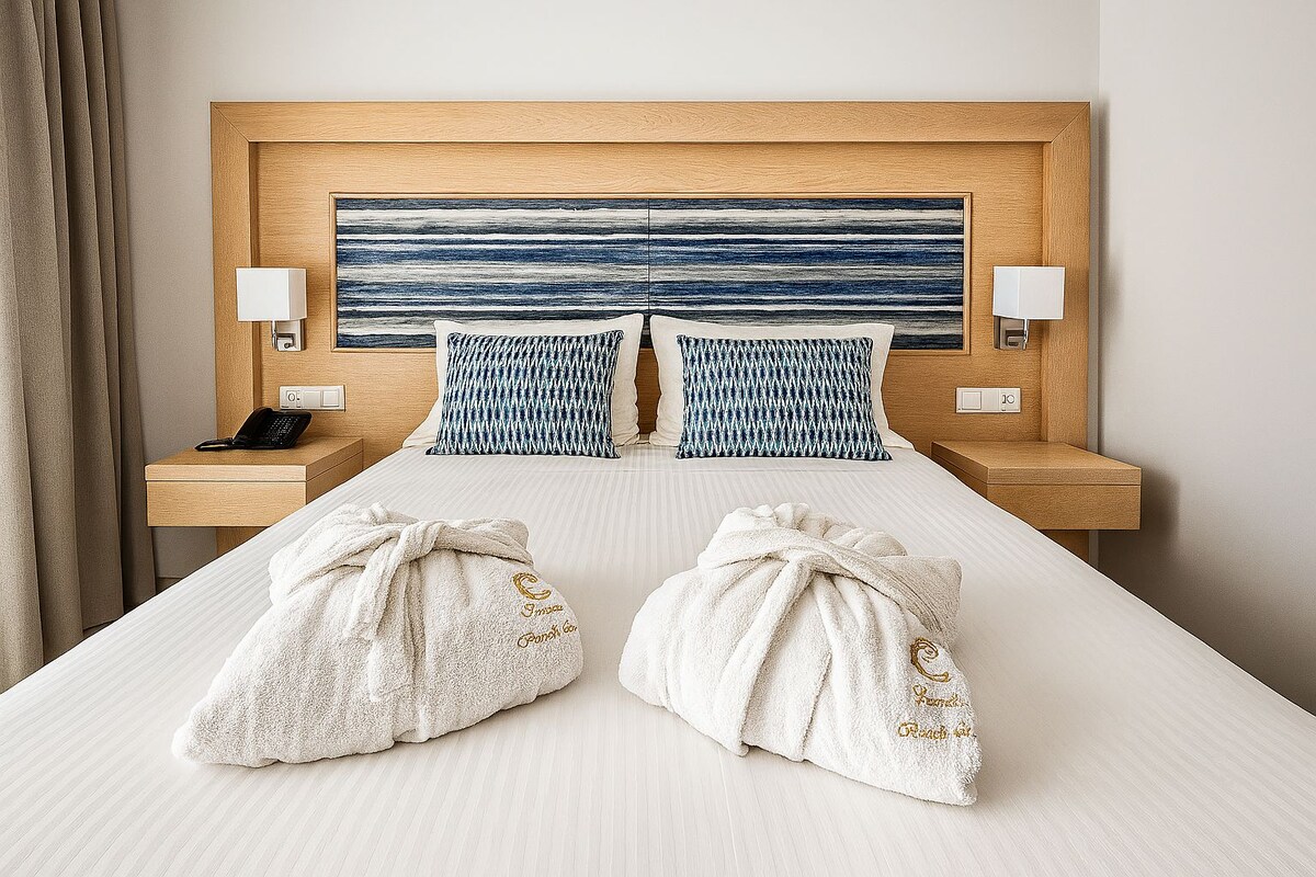 Habitación de hotel con una cama grande, dos lámparas de noche, una mesita de noche con un teléfono y dos toallas con texto bordado.