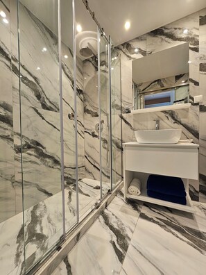 A modern bathroom with a glass-enclosed shower, marble walls, and a white vanity with a sink. - Paradise Resort Evia (Eretria)