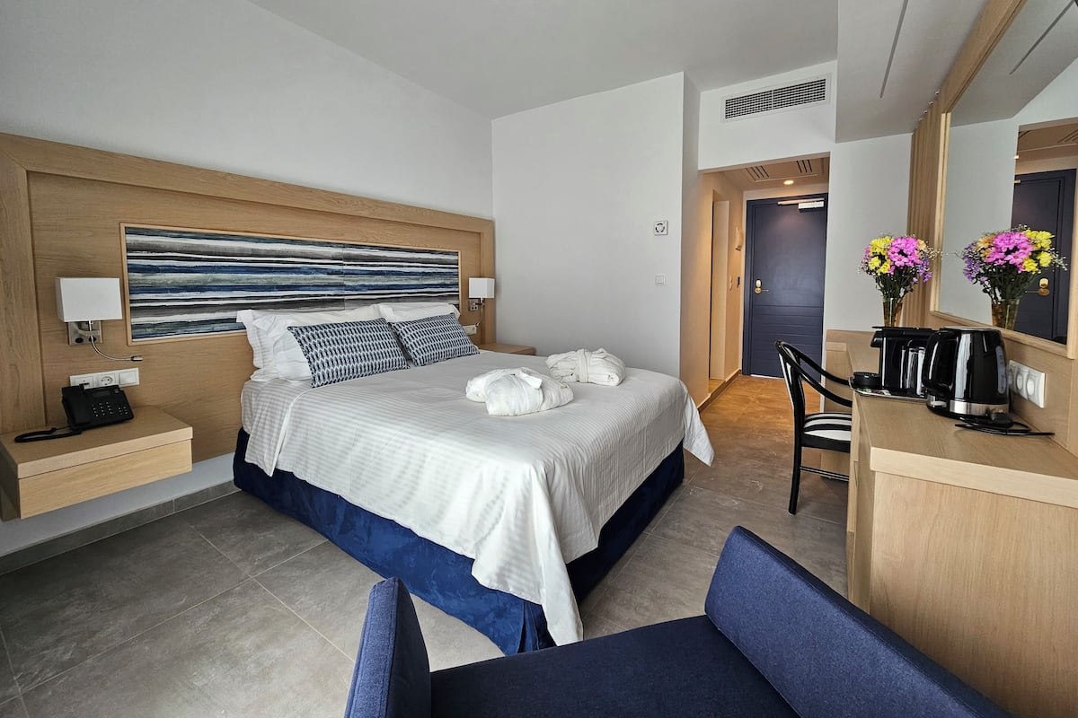A modern hotel room with a large bed, bedside tables, a desk with a chair, a coffee maker, and a mirror.