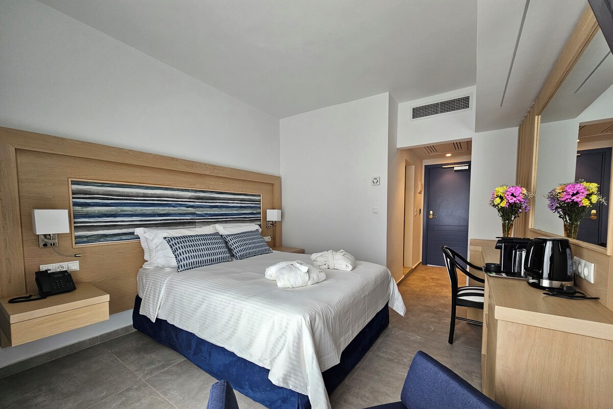 Una habitación de hotel moderna con una cama grande, mesitas de noche, un escritorio con silla, cafetera y espejo.