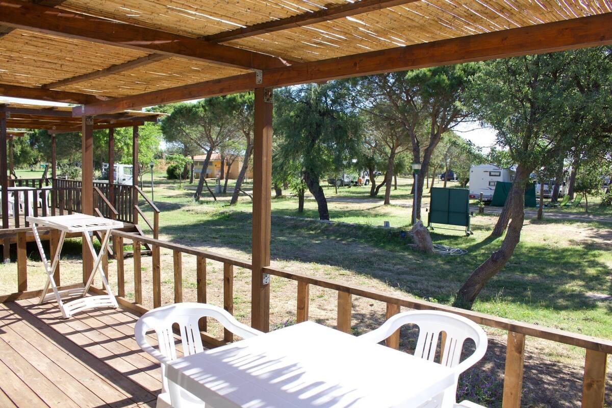 A covered outdoor area with wooden tables and chairs, a grassy lawn, and trees.