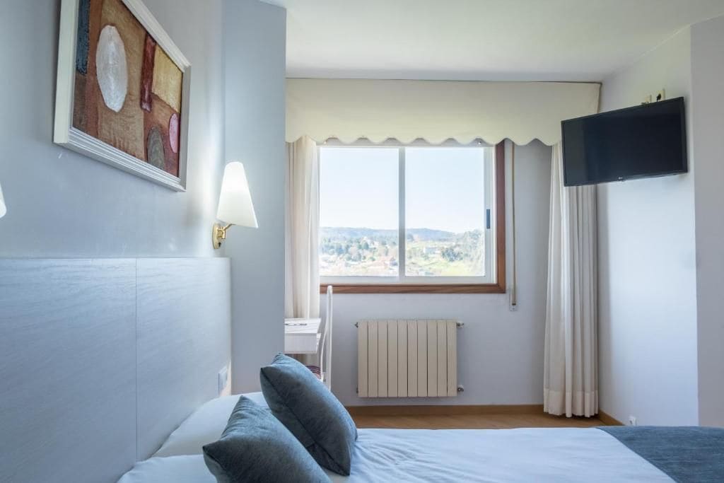 Un dormitorio con cama, ventana con cortinas, radiador, televisor montado en la pared y un cuadro.