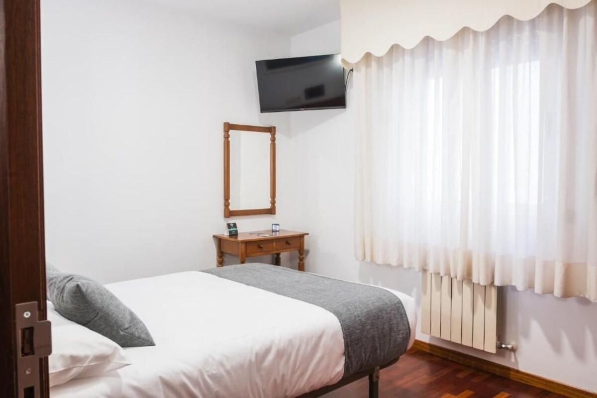 A hotel room with a bed, a wooden bedside table, a mirror, a television mounted on the wall, and a window with white curtains.