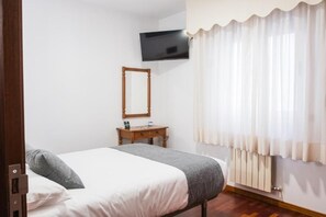 A hotel room with a bed, a wooden bedside table, a mirror, a television mounted on the wall, and a window with white curtains. - Alda Arteixo (Abegondo)
