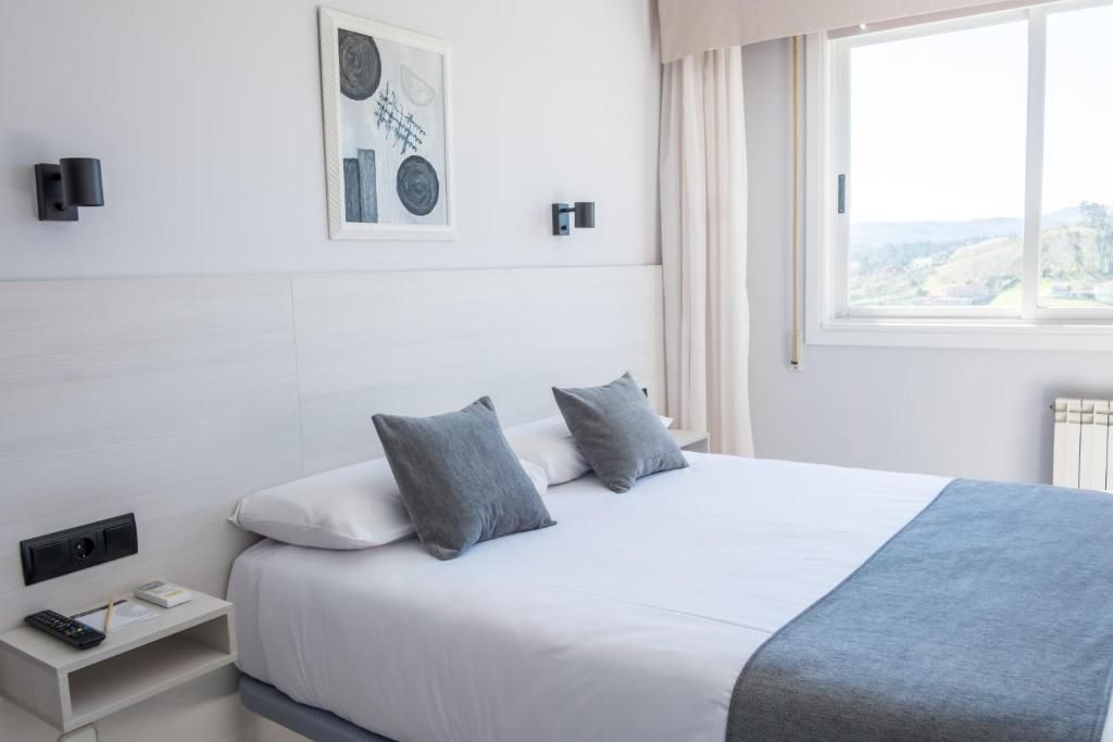 A modern hotel room with a large bed, a bedside table with a remote control, a framed artwork on the wall, and a window with a view.