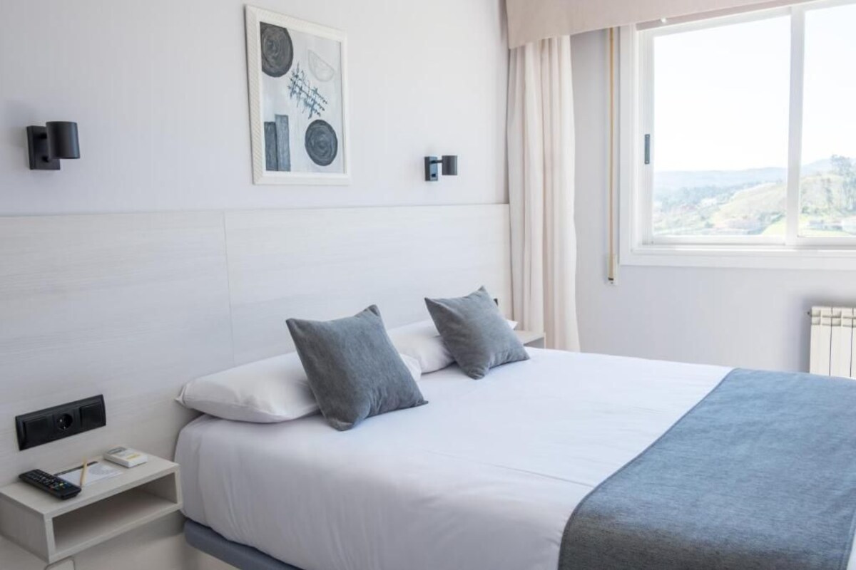 A modern hotel room with a large bed, a bedside table with a remote control, a framed artwork on the wall, and a window with a view.