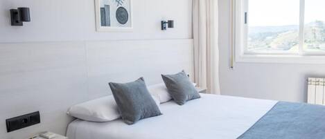 A modern hotel room with a large bed, a bedside table with a remote control, a framed artwork on the wall, and a window with a view.