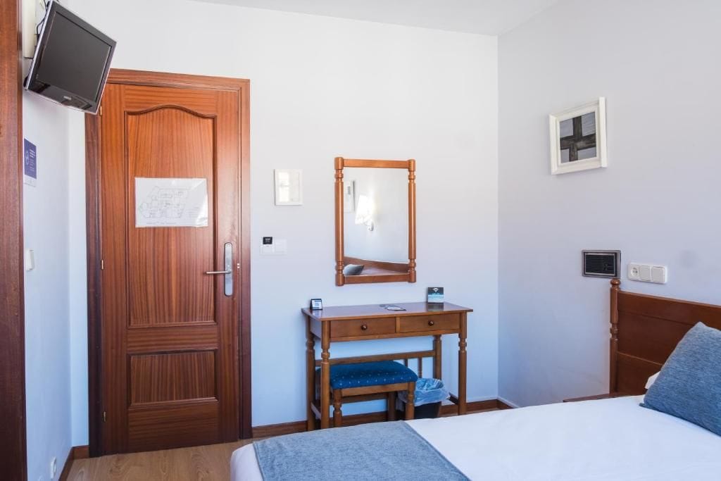 Habitación de hotel con puerta de madera, cama, mesita de noche con espejo, escritorio pequeño y televisor montado en la pared.
